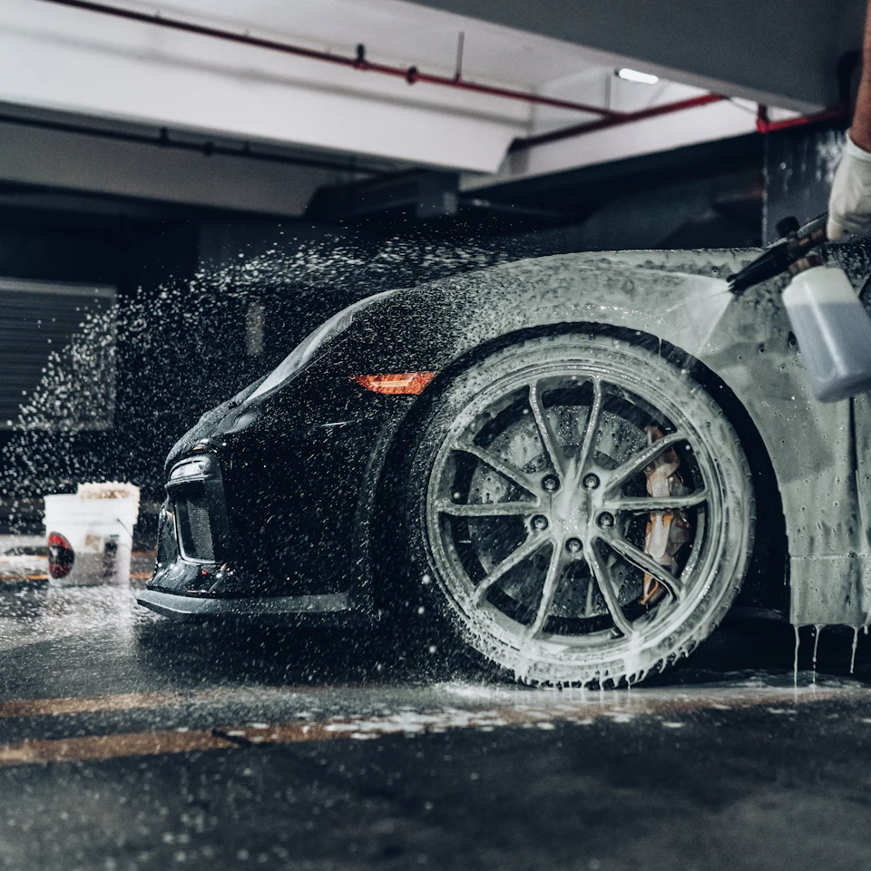 A person spraying soapy foam onto a black sports car in a garage
