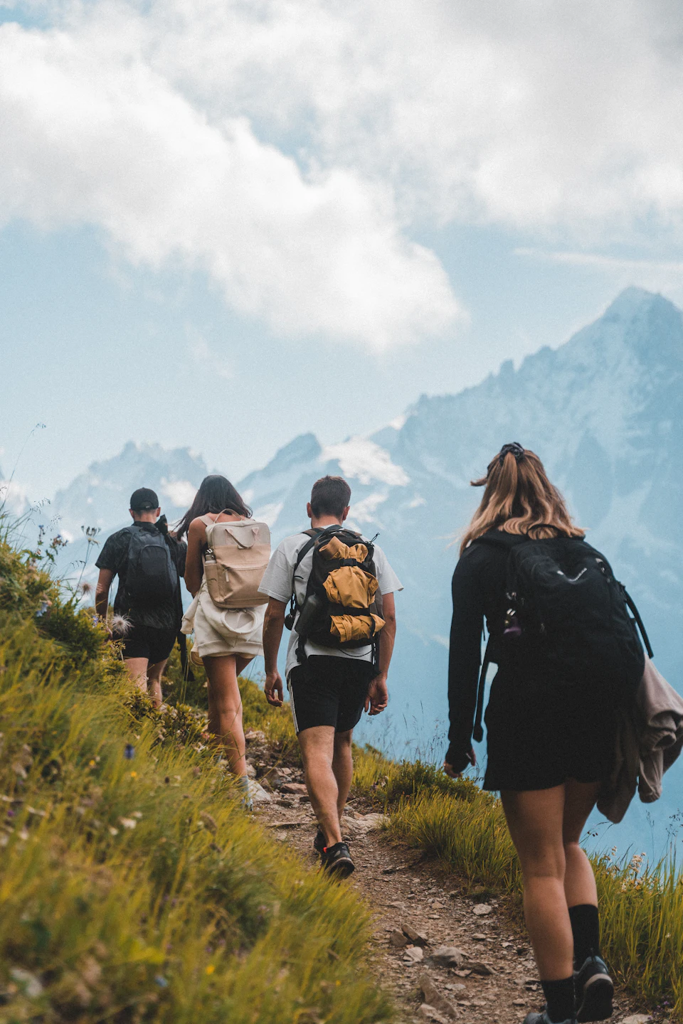 Four people with backpacks hiking on a grassy mountain trail toward snowy peaks