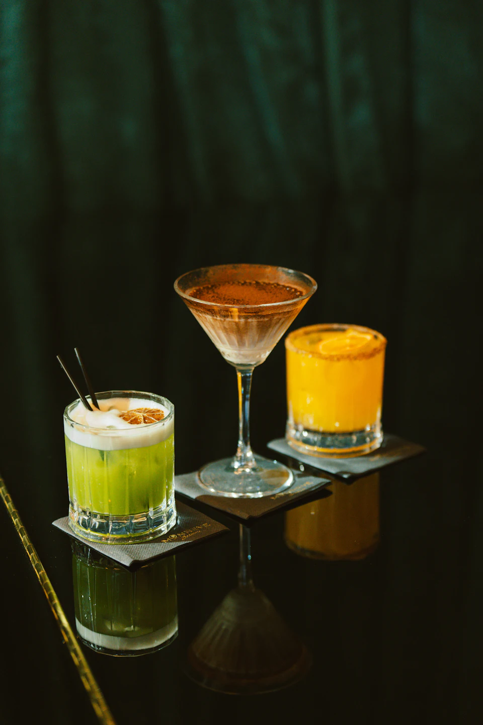 Three colorful cocktails arranged on a dark reflective surface
